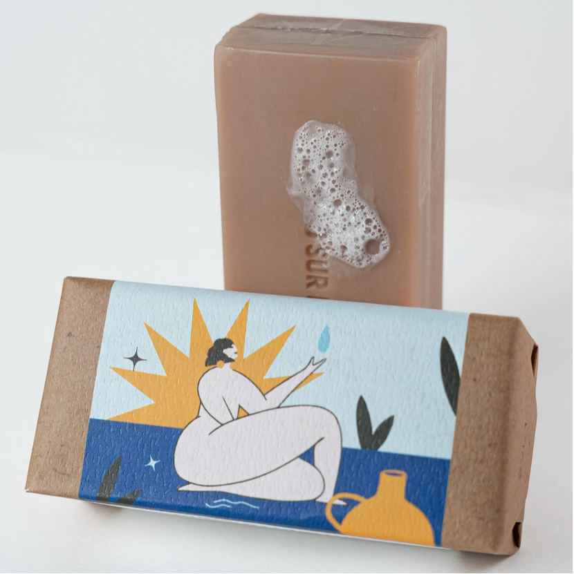 A rectangular bar of soap in a warm tan color stands upright behind its matching cardboard packaging. The box has a printed illustration of a seated figure in white, a yellow sunburst, blue water, and dark green leaves.