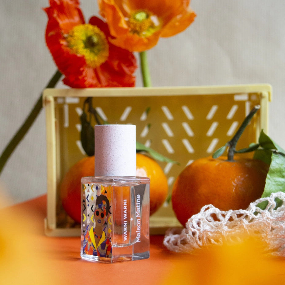 Small bottle of perfume with colorful label on a table with pumpkins and flowers.