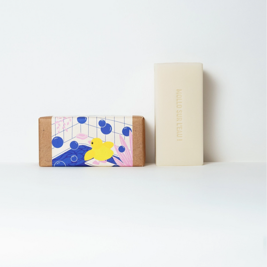Two rectangular bars of soap standing upright on a white surface against a white background. The left bar has a kraft paper border and a colorful illustration featuring a yellow duck, blue circles, and pink floral elements.