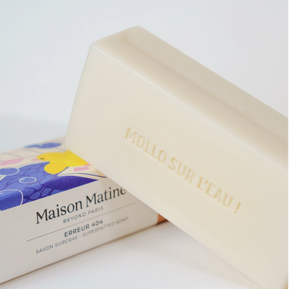 A close-up of a Maison Matiné bar of soap in a creamy off-white color, with the brand's colorful packaging box visible underneath. The bar of soap is embossed with the French phrase 'MOLLO SUR L'EAU !'.