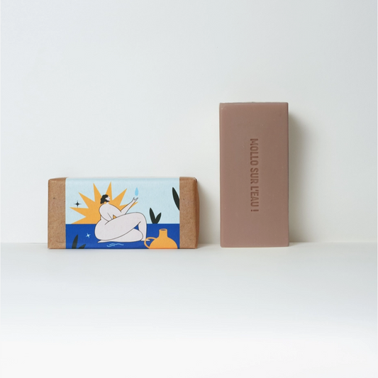 Two rectangular bars of soap on a white surface against a white background. The left bar has a colorful illustrated paper wrap showing a seated figure in a pool with a sun and blue water, and the right bar is plain taupe-brown with the vertical text 'MULO SUR KEALI'.