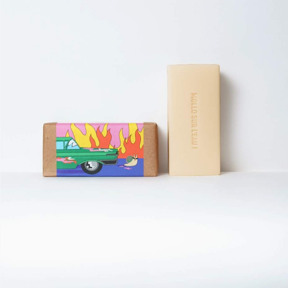 Two rectangular bars of soap standing upright on a white surface against a white background. The left bar is wrapped in kraft paper with a colorful illustration of a green car and flames in pink, yellow, and blue.