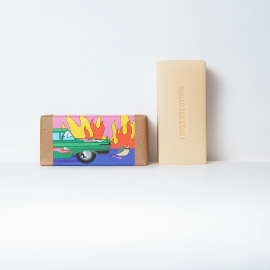 Two rectangular bars of soap standing upright on a white surface against a white background. The left bar is wrapped in kraft paper with a colorful illustration of a green car and flames in pink, yellow, and blue.