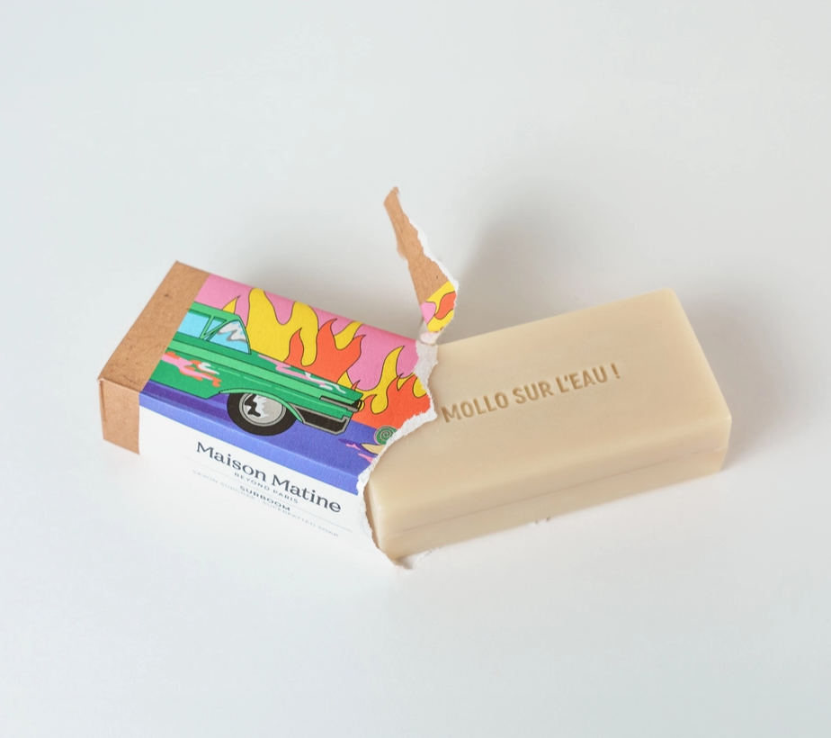 A beige bar of Maison Matine soap labeled 'MOLLO SUR L'EAU !' shown next to its colorful illustrated cardboard box on a clean white background.